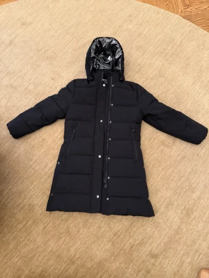 Svart lång dunjacka med huva - Snygg svart lång dunjacka med quiltad design och avtagbar huva. Jackan har dragkedja och tryckknappar framtill samt två fickor med dragkedja. Insidan har ett glansigt foder som ger extra värme och stil. Perfekt för kalla vinterdagar. Storlek 140.