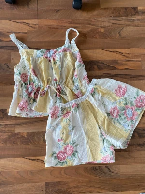 Blommig pyjamas-set med spetsdetaljer - Säljer ett somrigt pyjamas-set med linne och shorts i vitt, gult och pastellrosa. Setet har blommigt mönster, spetsband på axelbanden och dragsko i midjan på linnet. Materialet är tunt och krinklat, perfekt för varma nätter.