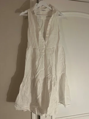 Vit ärmlös linneklänning från By Malina - Superfin vit kortklänning i linne från By Malina. Klänningen är ärmlös med v-ringning och knappar upptill, samt har volangdetaljer och en luftig passform. Perfekt för varma dagar och enkel att styla med accessoarer.