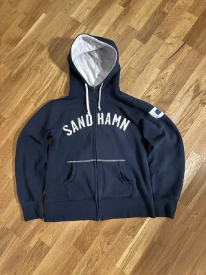 Marinblå Sandhamn zip-hoodie - Mörkblå hoodie med dragkedja från Sandhamn. Snyggt vitt tryck över bröstet och vit insida på huvan. Två fickor framtill och vita dragsnören i huvan. Liten blåvit patch på ena ärmen. Perfekt för bekväma outfits! Storlek ligger på S exakt