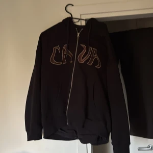 Svart hoodie från The Cava Company - Säljer en svart hoodie från The Cava Company med dragkedja och huva. På bröstet står det 'CAVA' i stora bokstäver. Hoodien har fickor framtill och justerbara snören i huvan. Perfekt för en avslappnad och cool stil.