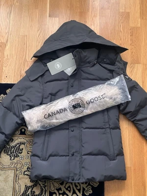 Canada Goose Wyndham-jacka i storlek S - Helt Ny - Denna Canada Goose Wyndham-jacka i storlek S är designad för både stil och värme. Med en klassisk grå färg och ett snölås som håller kylan ute, är den perfekt för kalla vinterdagar. Jackan levereras med ett fack för den borttagbara pälsen, vilket ger extra isolering. Med sina funktionella detaljer och den ikoniska loggan på ärmen, är denna jacka både ett praktiskt och lyxigt val för den som söker kvalitet och värme under vintern.