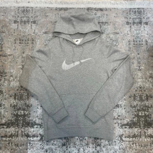Grå Nike hoodie med logga - Snygg grå hoodie från Nike med stor vit Swoosh-logga på bröstet. Klassisk modell med huva och dragsnören samt magficka framtill. Tillverkad i mjukt sweatshirtmaterial som är skönt att bära. Perfekt för en avslappnad och sportig stil.