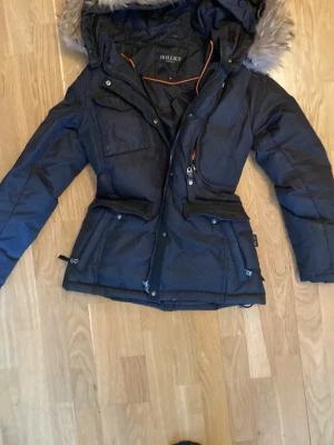 Svart dunjacka med fuskpäls från Hollies - Snygg svart dunjacka från Hollies med avtagbar huva och päls. Jackan har flera fickor med dragkedja, justerbar midja och detaljer i orange. Perfekt för kalla vinterdagar då den är riktigt varm.