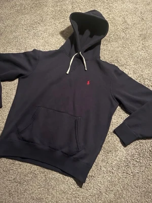 Mörkblå hoodie från Polo Ralph Lauren - Mörkblå hoodie med vit dragsko och klassisk röd Polo-logga broderad på bröstet. Stor känguruficka framtill och mjukt material som känns skönt mot huden. Perfekt för en avslappnad och stilren look.