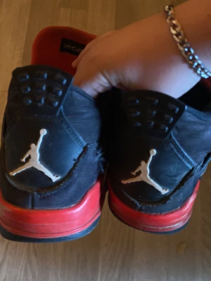 Svarta och röda Air Jordan 4 sneakers - Snygga Air Jordan 4 sneakers i svart med röda detaljer och klassisk Jumpman-logga på hälen. Skorna har en hög siluett och meshpaneler på sidorna samt plös. Perfekta för dig som gillar streetwear och sportig stil.