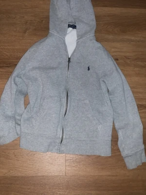 Grå hoodie från Polo Ralph Lauren - Klassisk grå hoodie med dragkedja från Polo Ralph Lauren. Hoodien har en broderad marinblå logga på bröstet, två fickor framtill och mjukt bomullsmaterial. Perfekt för en chill och stilren look. Strl 152