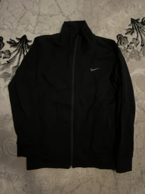 Svart träningsjacka från Nike - Snygg svart träningsjacka från Nike med hel dragkedja och hög krage. Jackan har diskret Nike-logga på bröstet och är tillverkad i ett lätt och stretchigt material som passar perfekt till träning eller uppvärmning.