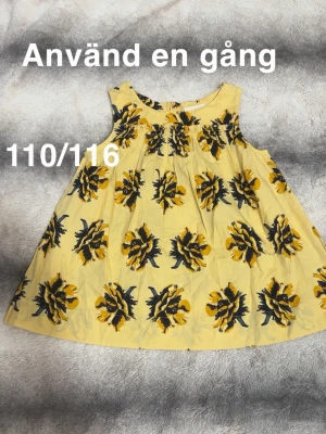 Gul blommig ärmlös linnetopp - Söt gul topp med stora blommiga mönster i svart och gult. Ärmlös modell med rund hals och luftig passform, perfekt för varma dagar. 🌸 Använd en gång. Storlek 110/116