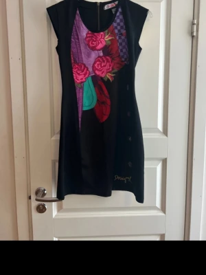 Svart  Desigual blommig - Unik svart kortklänning från Desigual med färgstarkt blommigt mönster i rosa, rött och lila framtill. Klänningen är ärmlös och har dragkedja i ryggen. Materialet känns mjukt och stretchigt, perfekt för dig som gillar statement-plagg