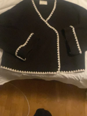 Svart cardiganjacka från Neo Noir - Svart cardiganjacka från Neo Noir med vita dekorativa kanter längs öppningen, fickorna och nederkanten. Jackan har en rak passform och två fickor framtill. Perfekt för lager-på-lager och snygg till jeans eller kjol.