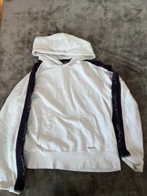 Vit hoodie från GANT med svarta detaljer - Snygg vit hoodie från GANT med svarta band längs ärmarna där det står GANT i vitt. Klassisk känguruficka framtill och broderad logga på magen. Mjuk bomullskänsla och ribbade muddar vid ärmslut och nederkant. Perfekt för en chill och stilren look.