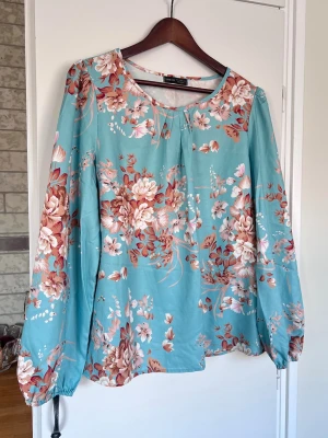 Blommig turkos blus, storlek L - Försvinner den 31:a mars om den inte sålts innan dess. Turkos blus med blommigt mönster i rostrött, vitt och brunt. Rund halsringning, långa ärmar med resår vid ärmslut och ett luftigt material. Använd 1 gång.