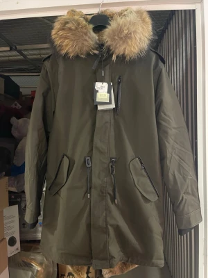 Olivgrön parkas från Mackage - Snygg olivgrön parkas från Mackage med stor pälskantad huva och flera dragkedjeförsedda fickor. Jackan har dragsko i midjan och detaljer i svart. Perfekt för kalla dagar och riktigt trendig look.