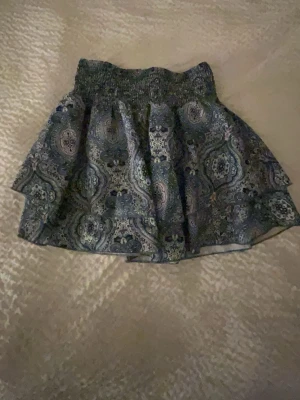 Mönstrad kortkjol med smockresår - Kortkjol med blått och grönt paisleyinspirerat mönster och bred smockresår i midjan. Kjolen har en lätt utställd form och är gjord i ett mjukt bomullsliknande material. Perfekt för dig som gillar färg och mönster!