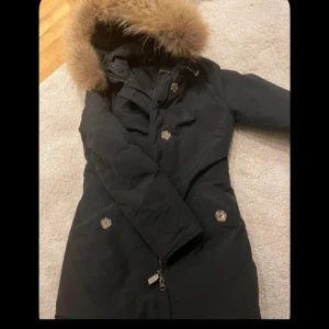 Svart parkas med päls - Svart parkas med lång passform och huva med fluffig päls. Jackan har stora knappar framtill, dragkedja och två snedställda fickor. Perfekt för kalla dagar och har en klassisk look som funkar till det mesta.