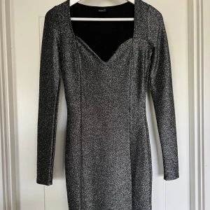Glittrig svart kortklänning - Svart glittrig klänning med lång ärm och figurnära passform. Klänningen har en v-ringad urringning. 