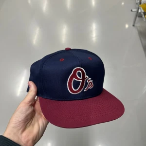 Blå och vinröd snapback keps O's - Snygg snapback keps i marinblått med vinröd skärm och broderad O's-logga framtill. Kepsen har justerbar baksida och grönt underskärm. Perfekt för dig som gillar sportig streetstyle och vill sticka ut med coola färger.