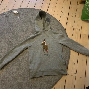 Grå hoodie Polo Bear Ralph Lauren - Säljer en grå hoodie från Ralph Lauren med Polo Bear-tryck på bröstet där björnen rider häst i röd-svart tröja. Klassisk huva med snörning och långa ärmar. Mjuk bomullsblandning och relaxed fit. Perfekt för dig som gillar streetwear och ikoniska prints. Finns et pytte litet ål i höger ficka men märks knappt 