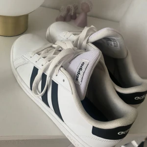 Adidas sneakers med svarta ”sträck” - Säljer ett par klassiska vita Adidas sneakers med svarta ränder på sidorna. Skorna har rund tå, platt sula och snörning framtill. Märket syns på både plös och häl. Materialet är syntet och textil, och designen är tidlös och clean.