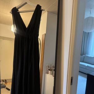 Svart långklänning med v-ringning - Elegant svart långklänning med djup v-ringning och ärmlös design. Klänningen har ett luftigt fall och markerad midja, perfekt för dig som gillar en stilren och tidlös look. Materialet ser ut att vara lätt och följsamt.