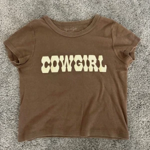 Brun cowgirl tröja - Brun croppad t-shirt med cowgirl-tryck i vitt framtill. T-shirten har korta ärmar, rund halsringning och är i mjuk bomull. 