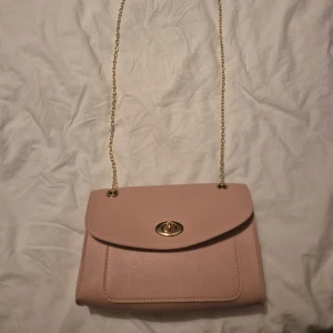 Ljusrosa axelväska med guldkedja - Söt ljusrosa axelväska i syntetmaterial med stilren design. Väskan har guldfärgad kedja som axelrem och ett snyggt vridlås i guld på framsidan. Perfekt storlek för det viktigaste och ger en chic touch till din outfit.