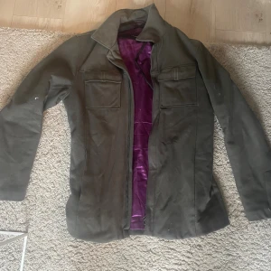 Mörkgrön jacka från J Lindeberg - Säljer en stilren mörkgrön jacka från J Lindeberg med lila satin-foder. Jackan har två bröstfickor med lock, klassisk krage och dragkedja framtill. Passar perfekt till dig som vill ha en clean look med lite edge.