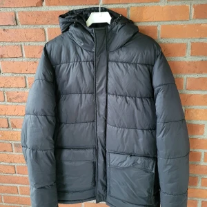 Svart pufferjack CUBUS stl L - Säljer en svart pufferjacka från Cubus i storlek L. Jackan har huva, stora fickor framtill och dragkedja. Den är vadderad och har ribbade muddar vid ärmsluten för extra komfort. Perfekt för kalla dagar när du vill hålla dig varm och snygg.