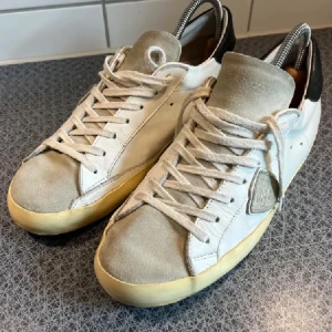 Philippe Model skor - Snygga vita sneakers från Philippe Model med beige mocka på tå och plös, svart häl och klassisk snörning. Skorna har en rund tå och platt sula, samt en ikonisk sköld på sidan. Perfekta för dig som gillar stilrena och trendiga sneakers. Använda få gånger, bara att höra av sig vid frågor och funderingar!