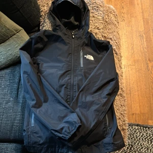 Svart regnjacka från The North Face - Svart regnjacka från The North Face med huva och dragkedja framtill. Jackan har meshfoder på insidan, två sidofickor med dragkedja och logga på bröstet och ryggen. Perfekt för blåsiga dagar och enkel att packa ner i väskan.
