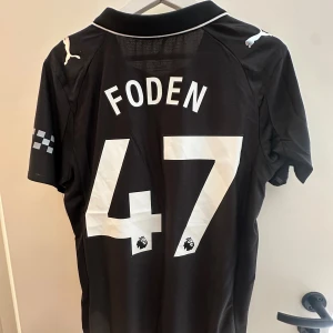 Foden Manchester City Bortaställ 25/26 - Säljer en svart Manchester City fotbollströja från Puma med Foden och nummer 47 på ryggen. Tröjan har korta ärmar, vit logga och detaljer, broderat klubbmärke och Etihad Airways-tryck på bröstet. Tillverkad i lätt och ventilerande funktionsmaterial.