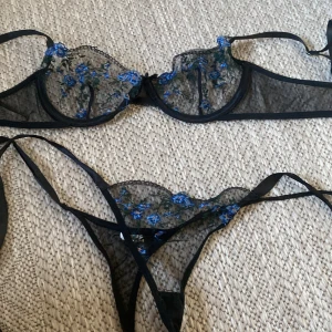 Svart blommig spetsunderklädeset - Säljer en svart korsett med transparent spets och blå broderade blommor. Toppen har tunna axelband och vacker broderad detalj framtill. Perfekt för dig som gillar romantisk och edgy stil.