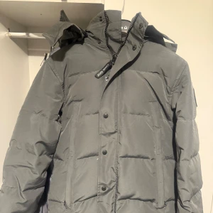 Canada Goose - Säljer en grå Canada Goose jacka i storlek S passar kanske även M. med huva och snygg patch på ärmen. Jackan har dragkedja och knappar framtill, samt två sidofickor. Perfekt för kalla dagar med sin tjocka, quiltade design och stilrena look. Pälsen och påsen följer med. 