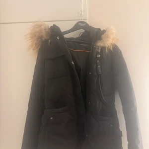 Svart parkas med päls från Hollies - Svart parkas från Hollies med fluffig beige päls på huvan. Jackan har flera fickor med knappar och dragkedja framtill. Snygg och varm modell med lång ärm och markerad midja. Perfekt för kalla dagar och har en klassisk look.