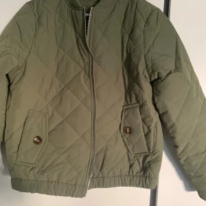 Olivgrön quiltad jacka med dragkedja - Snygg olivgrön quiltad jacka med dragkedja framtill och två fickor med knappar. Jackan har rund halsringning och elastisk nederkant för en skön passform. Perfekt för dig som gillar stilren och enkel design.