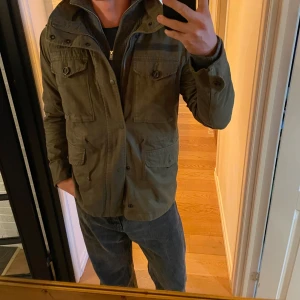 Olivgrön fieldjacka från Superdry - Tja! Säljer en olivgrön fieljacka från Superdry med flera fickor, knappar och dragkedja. Jackan har hög krage, axelklaffar och coola detaljer som ger en urban vibe. Perfekt för dig som gillar streetstyle och funktionella plagg samt passar in nu mot hösten. Hör av dig vid intresse!