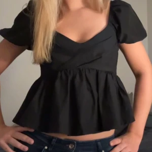 Svart blus med puffärm och v-ringning - Supersnygg svart blus med puffiga korta ärmar och v-ringad omlott framtill. Modellen har en markerad midja och volangig nederdel som ger en feminin siluett. Perfekt att styla med jeans eller kjol för en trendig look.