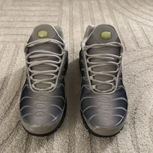 Nike Air Max Plus Tn grå/svarta sneakers - Nike Air Max Plus Tn sneakers i grått och svart med vita detaljer och gula inslag. Ovandel i mesh och syntet, ikoniska vågformade linjer och Tn-logga bak. Skorna har snörning och en chunky svart sula med synliga Air-enheter för extra komfort. Har för många skor nu och vill bli av med några 
