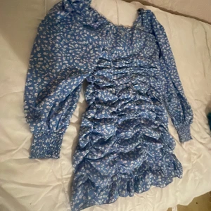 Blå blommig volangklänning med långa ärmar - Supersöt blå kortklänning med vitt blommönster och volanger framtill. Klänningen har långa puffärmar med smock vid ärmslut och dragkedja i sidan. Den ör mer somrigt hade på mig under midsommaren en gång bara