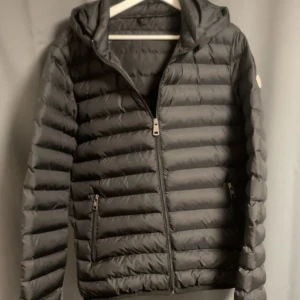 Svart Moncler dunjacka med huva - Svart dunjacka från Moncler med huva och klassisk logga på ärmen. Jackan har horisontella sömmar, två fickor med dragkedja och en smidig dragkedja framtill. Perfekt för kyliga dagar och riktigt snygg passform. Passar L också