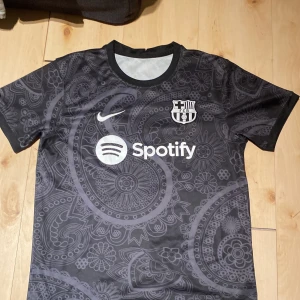 Svart Barcelona Nike fotbollströja - Snygg svart Barcelona fotbollströja från Nike med Spotify-logga på bröstet. Tröjan har ett unikt paisleymönster i mörkgrå toner och klassisk rund hals. Tillverkad i lätt och ventilerande material, perfekt för match eller träning.