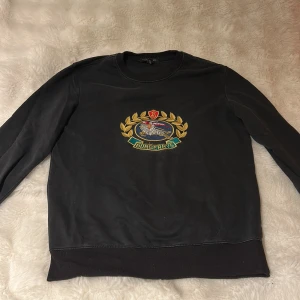 Svart Burberrys sweatshirt med brodyr - Svart sweatshirt från Burberrys med ikonisk broderad logga i guld, rött och blått på bröstet. Tröjan har rund hals, ribbade muddar och är tillverkad i mjuk bomull. Perfekt för dig som vill ha en klassisk och stilren look med lyxig känsla.