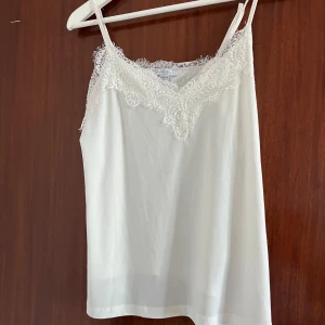 Vit linnetopp med spets från Amisu - Säljer en vit linnetopp från Amisu i storlek S. Toppen har tunna axelband och v-ringning med fina spetsdetaljer längs kanten. Perfekt att styla med jeans eller kjol för en fräsch look.