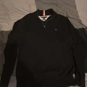Svart half zip-tröja Tommy Hilfiger - Svart långärmad tröja från Tommy Hilfiger i premium bomull. Klassisk half zip med ståkrage och diskret logga på bröstet. Kragen har snygga ränder i rött och vitt som ger en sportig touch. Perfekt för lager-på-lager och stilren vardagslook.