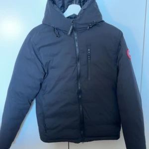 Svart dunjacka från Canada Goose - Svart dunjacka från Canada Goose med huva och dragkedja framtill. Jackan har en klassisk patch på ärmen, flera fickor med dragkedja och en stilren, sportig look. Perfekt för kalla dagar och riktigt snygg till streetwear. Bra skick men finns rtt litet märke från ett öagat hål, de är dock inget man ser, kan även gå med på byten!! På nått i ungefär samma värde