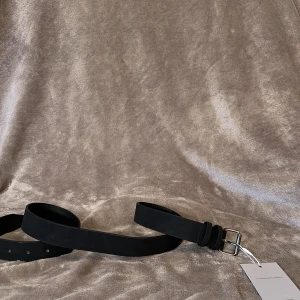 Svart bälte - Stilrent svart bälte med matt silverfärgat spänne. Bältet har en klassisk, rak form och är gjort i skinn med ljusbrun insida. Perfekt accessoar för att lyfta din outfit. 100cm