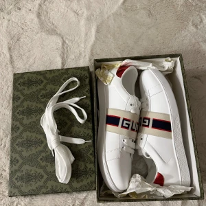 Gucci skor - Säljer ett par vita Gucci sneakers i skinn med breda elastiska band över foten med GUCCI-logga i rött och blått. Skorna har snörning, rund tå och röd metallic-detalj på hälen. Snygg och stilren design med platt sula och exklusiv präglad undersida. Helt nya från butik kostar. Slutsålda i butik. Enda som finns i marknaden är använda. Dem här är helt nya. Med box och allt. Först till kvarn.