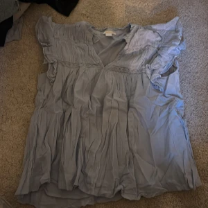 Ljusblå blus med volang från Gina Tricot - Säljer en ljusblå blus från Gina Tricot i storlek 170. Blusen har v-ringad hals, korta volangärmar och dekorativ spetsdetalj under bysten. Materialet är mjukt och luftigt, perfekt för vår och sommar. Plagget har en loose passform och ett romantiskt uttryck.