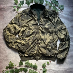 Woolrich Reversible Bomber Jacket – Storlek S - Exklusiv vändbar bomberjacka från Woolrich, där du får två stilar i en – klassisk militärgrön på ena sidan och modern camouflage på den andra.  🎨 Färg: Militärgrön / Camouflage 📏 Storlek: S 📐 Mått: Fråga ifall det önskas! ♻️ Skick: Mycket bra – knappt använd, inga defekter 🧵 Material: Polyamid / Bomull